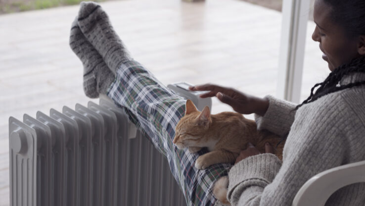 Frau streichelt eine Katze und sitzt in der Winterregenzeit bequem mit Beinen auf dem Heizkörper zu Hause vor dem großen Fenster. Die Frau trägt Wollsocken und benutzt bei kaltem Wetter eine Heizung.
