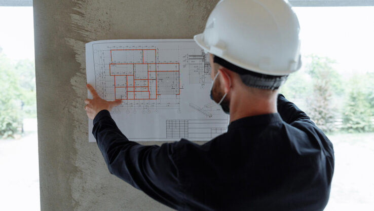 Mann steht mit einem Schutzhelm in einem Neubau und schaut sich den Bauplan an