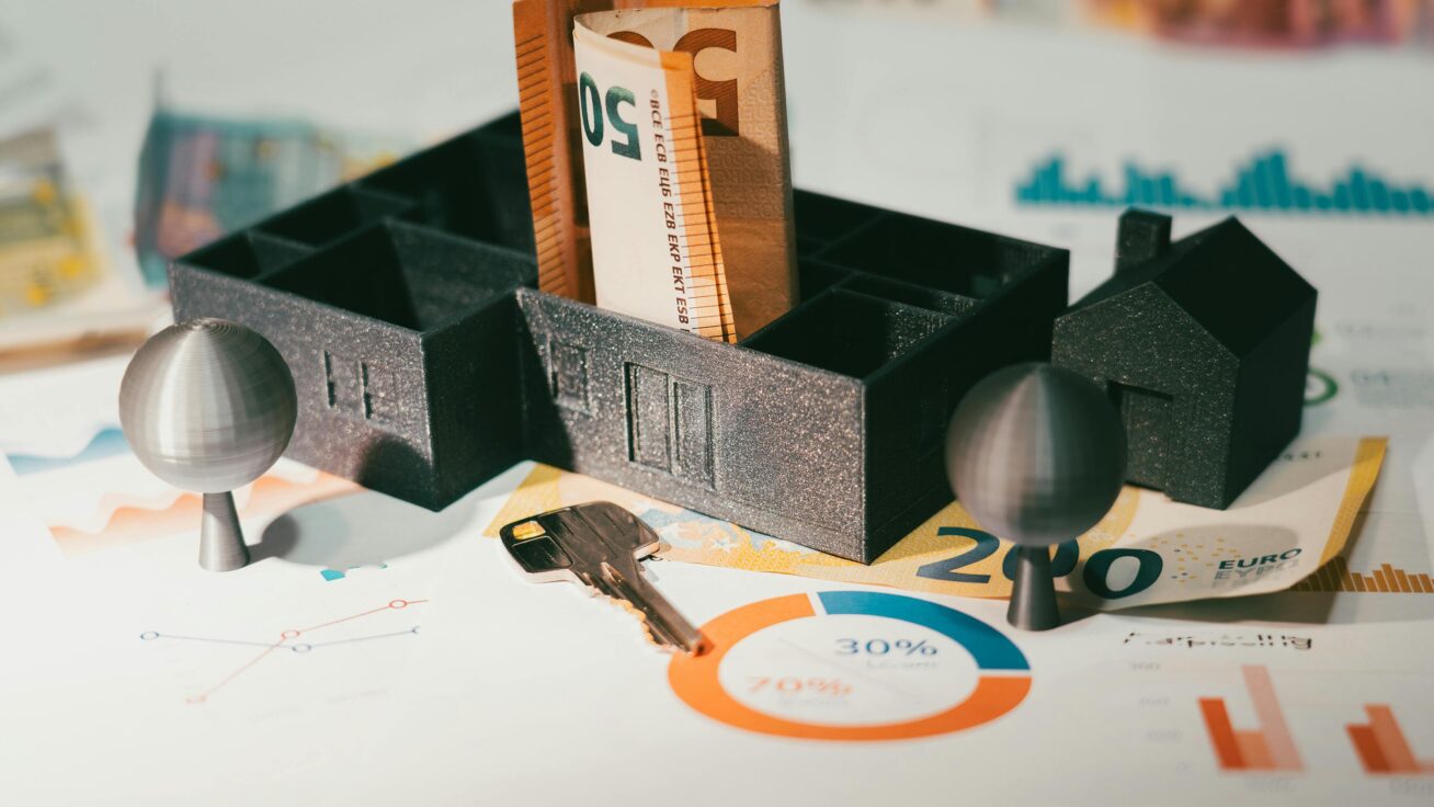 Modelhaus auf dem Geldscheine liegen. Ein Schlüssel liegt vor dem Modellhaus und es steht auf einem Plan.