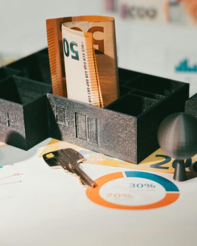 Modelhaus auf dem Geldscheine liegen. Ein Schlüssel liegt vor dem Modellhaus und es steht auf einem Plan.